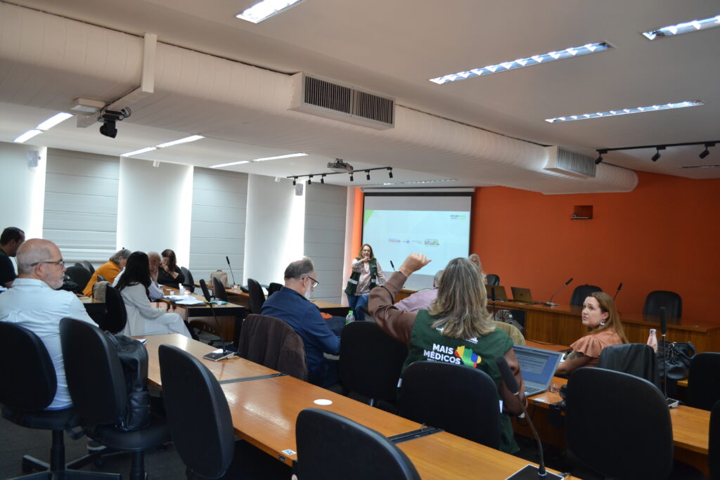 Participantes da reunião do Programa Mais Médicos na FCM acompanham uma apresentação projetada. Uma mulher em pé fala ao microfone enquanto outras pessoas interagem e fazem anotações. Um dos participantes levanta a mão.