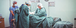 Equipe médica realiza procedimento cirúrgico em uma sala de cirurgia. Quatro profissionais de saúde, usando roupas cirúrgicas, máscaras e toucas, estão concentrados ao redor do paciente deitado na maca. Equipamentos médicos, suporte de soro e bandeja com instrumentos cirúrgicos compõem o ambiente estéril da sala.