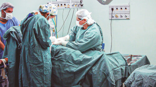 Equipe médica realiza procedimento cirúrgico em uma sala de cirurgia. Quatro profissionais de saúde, usando roupas cirúrgicas, máscaras e toucas, estão concentrados ao redor do paciente deitado na maca. Equipamentos médicos, suporte de soro e bandeja com instrumentos cirúrgicos compõem o ambiente estéril da sala.