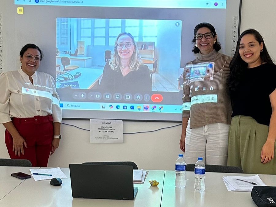 Três mulheres posam para foto em uma sala de aula, duas de pé ao lado direito e esquerdo da tela de projeção, e a terceira participando por videoconferência projetada na tela. À frente, sobre a mesa, há um notebook, papéis e garrafas de água.