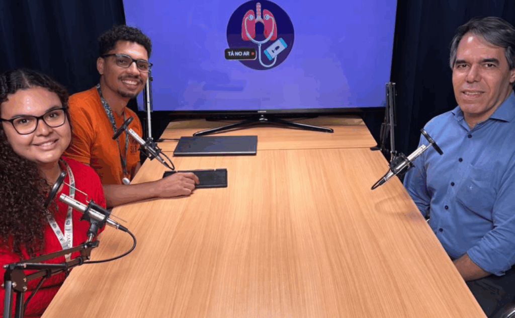 Três pessoas estão sentadas ao redor de uma mesa de madeira clara, em um estúdio de gravação de podcast. À esquerda, uma jovem de cabelo cacheado, longo e escuro, veste blusa vermelha e usa óculos de grau. Ao lado dela, no centro, está um homem de pele morena, com cabelo curto e barba rala. Ele veste uma camiseta laranja e também usa óculos de grau. À direita, está um homem de cabelo grisalho e pele clara, vestindo uma camisa azul. Todos estão sorrindo em direção à câmera. À frente de cada pessoa há um microfone preso em suportes articulados. Ao fundo, há uma televisão com fundo azul que exibe o logotipo do podcast “Tá no Ar”, composto por uma ilustração de pulmões com um estetoscópio ligado a um celular, simbolizando a relação entre saúde e comunicação.