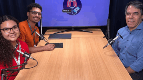 Três pessoas estão sentadas ao redor de uma mesa de madeira clara, em um estúdio de gravação de podcast. À esquerda, uma jovem de cabelo cacheado, longo e escuro, veste blusa vermelha e usa óculos de grau. Ao lado dela, no centro, está um homem de pele morena, com cabelo curto e barba rala. Ele veste uma camiseta laranja e também usa óculos de grau. À direita, está um homem de cabelo grisalho e pele clara, vestindo uma camisa azul. Todos estão sorrindo em direção à câmera. À frente de cada pessoa há um microfone preso em suportes articulados. Ao fundo, há uma televisão com fundo azul que exibe o logotipo do podcast “Tá no Ar”, composto por uma ilustração de pulmões com um estetoscópio ligado a um celular, simbolizando a relação entre saúde e comunicação.