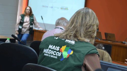 Reunião do Programa Mais Médicos realizada em uma sala de conferências, com foco na ampliação e qualificação da Atenção Básica. Participantes usam coletes verdes com o logo do programa e estão voltados para uma apresentação projetada na parede.