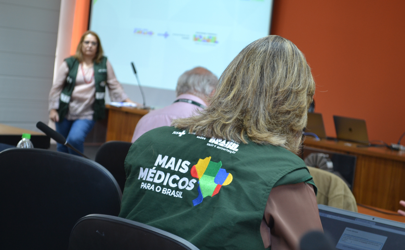 Reunião do Programa Mais Médicos realizada em uma sala de conferências, com foco na ampliação e qualificação da Atenção Básica. Participantes usam coletes verdes com o logo do programa e estão voltados para uma apresentação projetada na parede.