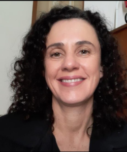 Mulher de cabelos cacheados e escuros, vestindo roupa preta, sorrindo para a câmera em um ambiente interno com móveis de madeira ao fundo.