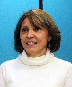 Mulher de cabelos curtos castanhos com mechas claras, vestindo blusa de gola alta branca, em frente a um fundo azul. Ela está olhando ligeiramente para o lado e sorrindo.