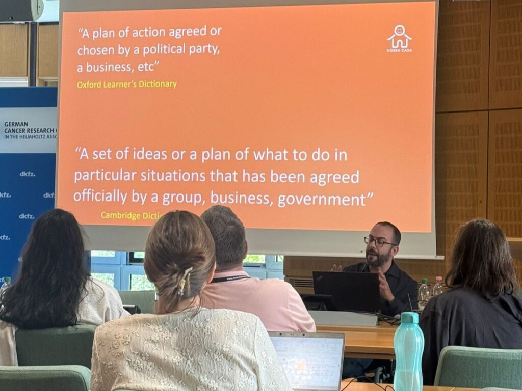 Homem usando óculos e camisa preta conduz oficina em sala de conferências, falando para um grupo sentado de frente para ele. Ao fundo, projeção de slide com definições de “plano de ação” dos dicionários Oxford e Cambridge, com fundo laranja.