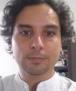 Homem com cabelo castanho ondulado, pele clara e barba rala, usando camisa branca com gola estilo mandarim, olhando diretamente para a câmera em um ambiente interno com prateleira ao fundo.