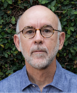 Homem calvo com barba e bigode grisalhos, usando óculos redondos e camisa azul, posando sério para a câmera diante de uma parede coberta por folhagem verde.