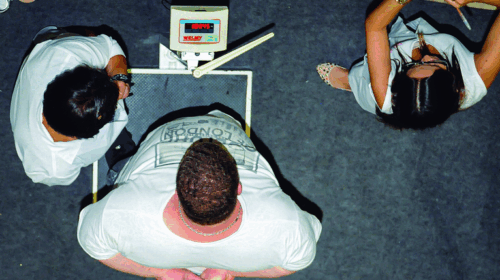 Imagem vista de cima mostrando duas pessoas, uma delas sobre uma balança digital que marca 169,4 kg, enquanto outra observa. Ao lado, uma terceira pessoa, vestindo roupas claras, anota informações em uma prancheta.