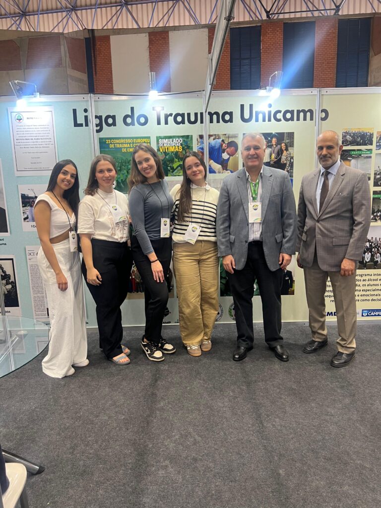Grupo de seis pessoas no estande da Liga do Trauma da Unicamp durante o congresso WCTEL. À esquerda, quatro estudantes da Liga, ao centro o professor Gustavo Fraga, e à direita o professor Hassan Al-Thani, todos sorrindo para a foto.