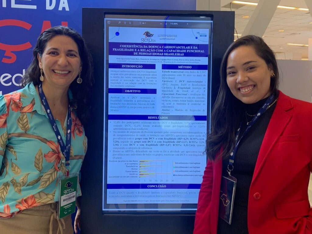 Duas mulheres sorridentes posam ao lado de um painel digital que exibe um pôster científico intitulado “Coexistência da doença cardiovascular e da fragilidade e relação com a capacidade funcional de pessoas brasileiras”. O pôster apresenta introdução, objetivo, método, resultados, conclusão e um gráfico, com logos institucionais na parte superior.