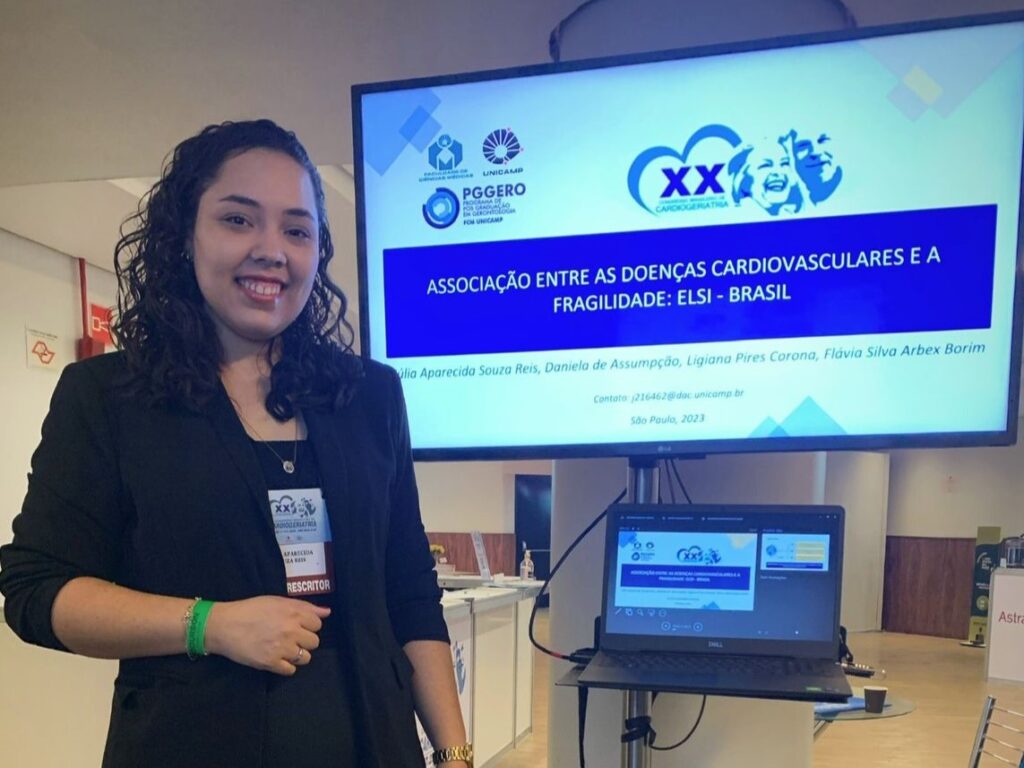 Mulher sorridente, usando blazer preto, posa ao lado de um monitor que exibe apresentação intitulada “Associação entre as doenças cardiovasculares e a fragilidade: ELSI – Brasil”, com nomes das autoras e logos institucionais.