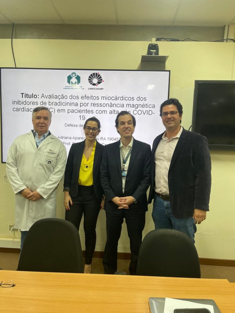 Quatro pessoas posam sorridentes em frente a uma tela de apresentação na Unicamp. A tela mostra o título de um trabalho acadêmico: “Avaliação dos efeitos miocárdicos dos inibidores de bradicinina por ressonância magnética cardíaca (RMC) em pacientes com alta de COVID-19”. À esquerda, um homem usa jaleco branco. Ao lado, uma mulher veste blazer preto e blusa amarela. À direita dela, um homem de terno escuro e crachá, e outro homem com blazer preto, camisa listrada e jeans.