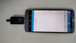 Telefone celular conectado a um dispositivo USB externo preto com luz indicadora azul, exibindo na tela um aplicativo com gráfico vazio.