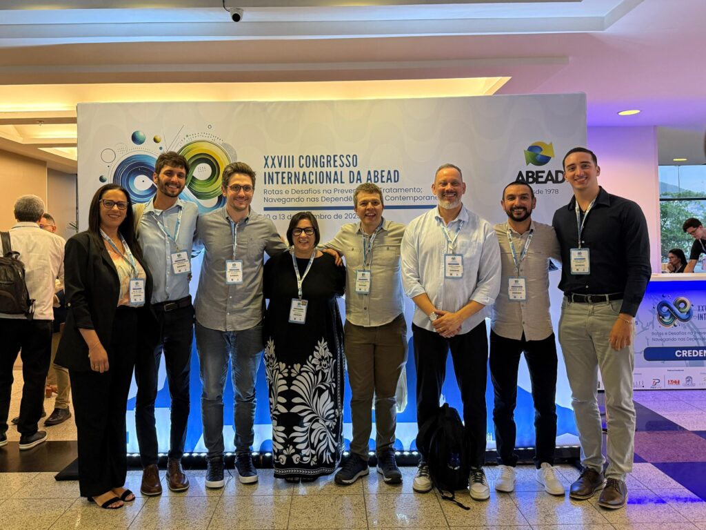 Um grupo de oito pessoas posando em frente ao painel do XXVIII Congresso Internacional da ABEAD. Todos usam crachás de identificação e sorriem para a foto. O painel traz o logotipo da ABEAD e a frase “Rotas e Desafios na Prevenção e Tratamento; Navegando nas Dependências da Contemporaneidade”.