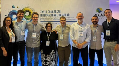 Um grupo de oito pessoas posando em frente ao painel do XXVIII Congresso Internacional da ABEAD. Todos usam crachás de identificação e sorriem para a foto. O painel traz o logotipo da ABEAD e a frase “Rotas e Desafios na Prevenção e Tratamento; Navegando nas Dependências da Contemporaneidade”.