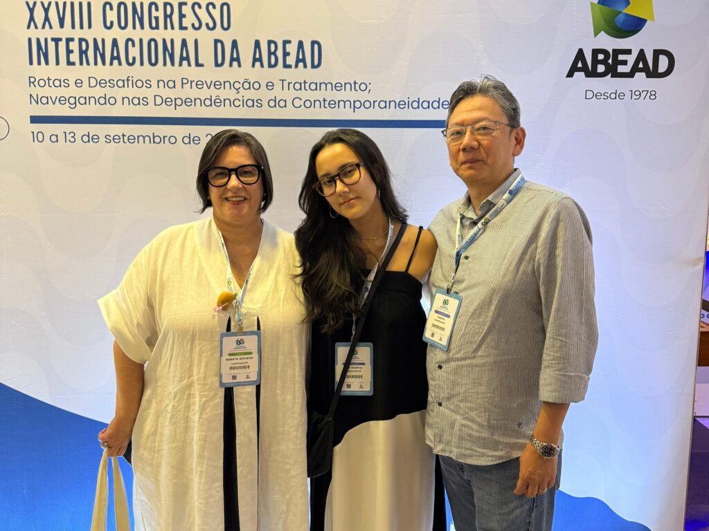 Três pessoas posam lado a lado em frente ao painel do XXVIII Congresso Internacional da ABEAD. Duas mulheres estão à esquerda e ao centro, e um homem à direita. Todos usam crachás de identificação do evento.