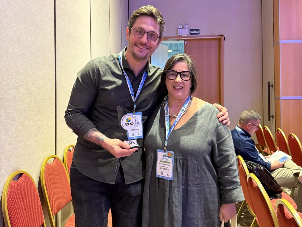 Duas pessoas posam juntas em um auditório durante o XXVIII Congresso Internacional da ABEAD. O homem segura um troféu e sorri, enquanto abraça a mulher ao lado dele, que também sorri para a câmera. Ambos usam crachás de identificação.