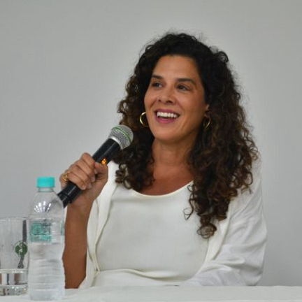 Mulher sorridente, de cabelos cacheados, usando blusa branca e segurando microfone, sentada à mesa durante um evento