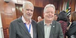 Dois homens posam sorridentes em um auditório. Ambos vestem ternos escuros — o da esquerda usa camisa rosa clara e medalha pendurada no pescoço, enquanto o da direita veste camisa cinza clara. Ao fundo, outras pessoas conversam em pé, e há bandeiras do Brasil e do Estado de São Paulo. O ambiente tem cadeiras azuis e paredes de madeira.