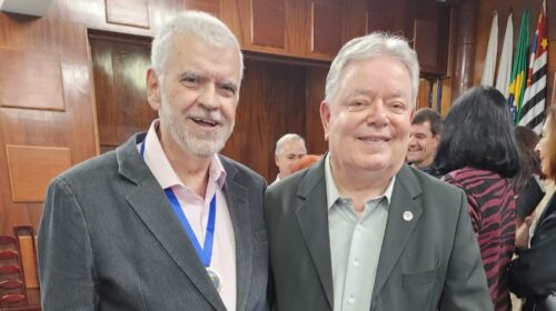 Dois homens posam sorridentes em um auditório. Ambos vestem ternos escuros — o da esquerda usa camisa rosa clara e medalha pendurada no pescoço, enquanto o da direita veste camisa cinza clara. Ao fundo, outras pessoas conversam em pé, e há bandeiras do Brasil e do Estado de São Paulo. O ambiente tem cadeiras azuis e paredes de madeira.