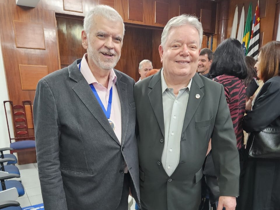 Dois homens posam sorridentes em um auditório. Ambos vestem ternos escuros — o da esquerda usa camisa rosa clara e medalha pendurada no pescoço, enquanto o da direita veste camisa cinza clara. Ao fundo, outras pessoas conversam em pé, e há bandeiras do Brasil e do Estado de São Paulo. O ambiente tem cadeiras azuis e paredes de madeira.