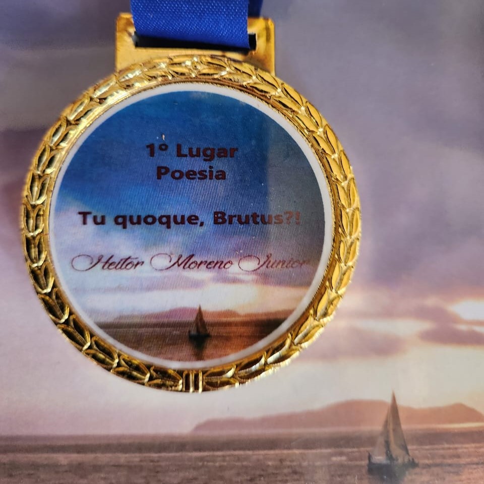 Medalha dourada com fita azul, exibindo no centro a inscrição: “1º Lugar Poesia – Tu quoque, Brutus?! – Heitor Moreno Júnior”. O fundo do medalhão mostra a imagem de um pôr do sol sobre o mar, com um veleiro em destaque.