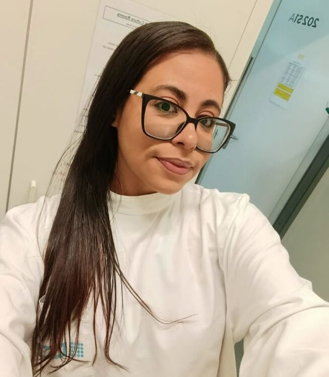 Pesquisadora jovem, de cabelos lisos e longos, usando óculos de armação preta e jaleco branco, em ambiente de laboratório, com portas e armários ao fundo.