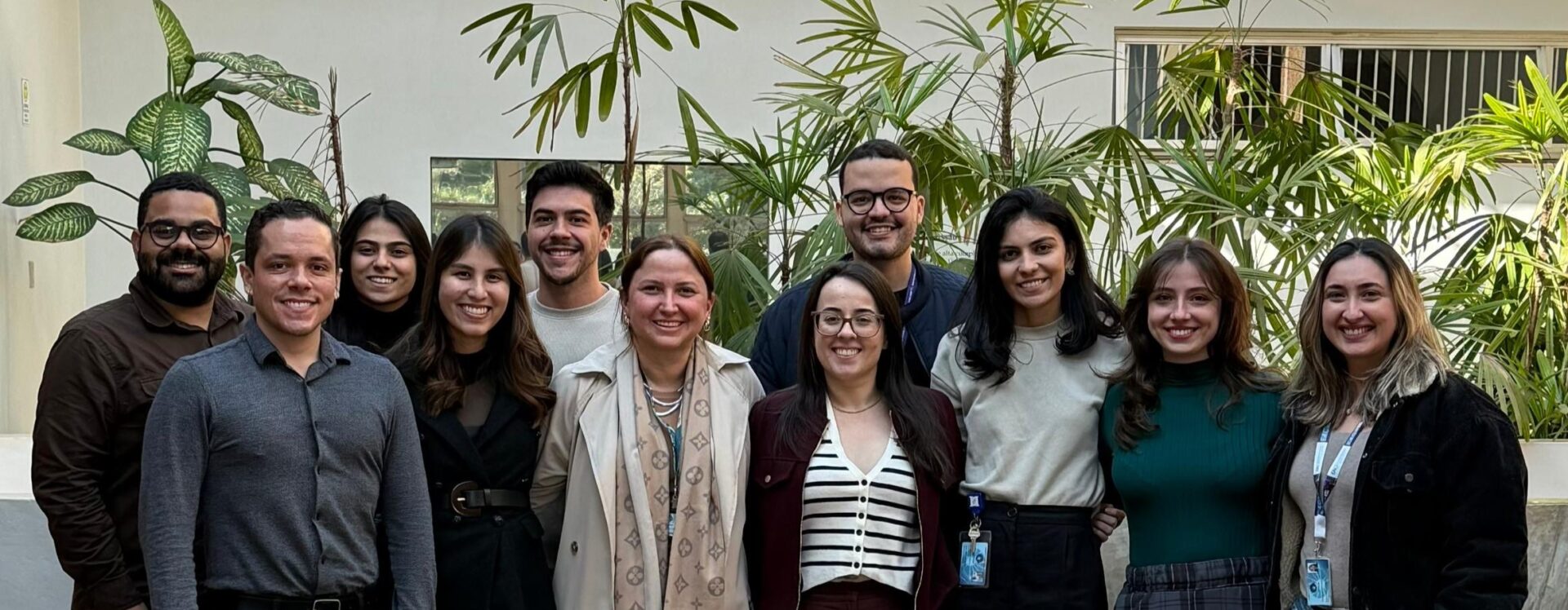 Um grupo de onze pessoas em pé, lado a lado, sorrindo para a câmera em ambiente interno com plantas ao fundo.