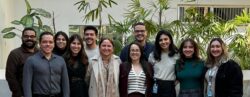 Um grupo de onze pessoas em pé, lado a lado, sorrindo para a câmera em ambiente interno com plantas ao fundo.