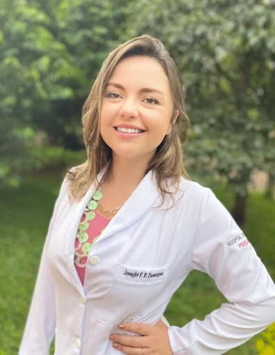 Mulher sorridente, de cabelos lisos, usando jaleco branco, em ambiente externo com vegetação ao fundo.