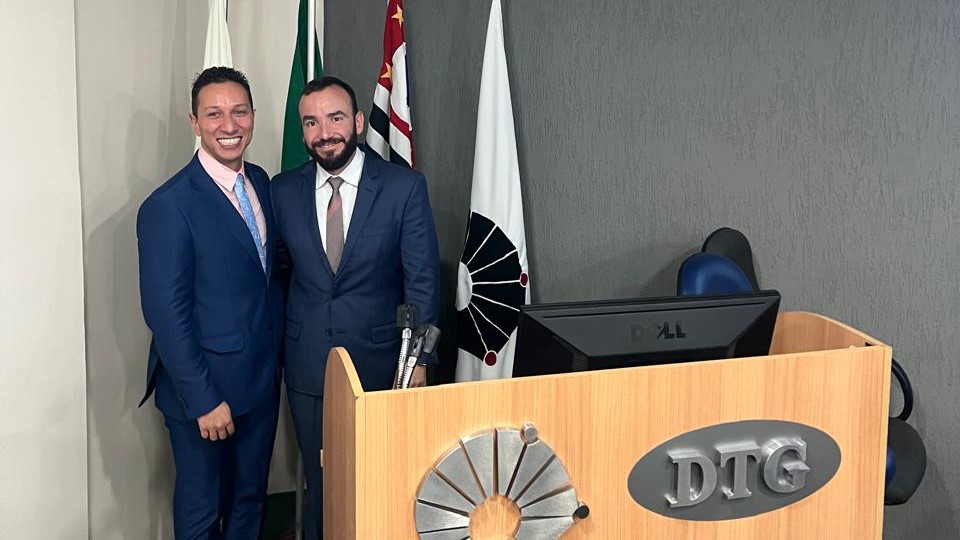 Duas pessoas de terno posam sorridentes atrás de um púlpito de madeira com a sigla "DTG". Atrás deles estão bandeiras do Brasil e do estado de São Paulo.