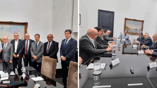 "Reunião institucional com autoridades e representantes, realizada em sala de paredes brancas com mesa de reuniões de vidro preto. Na primeira foto, o grupo de participantes posa em pé para registro, lado a lado, diante de uma pintura emoldurada. Na segunda foto, as mesmas pessoas aparecem sentadas ao redor da mesa, em discussão, com copos de água, papéis e crachás de identificação sobre a mesa."