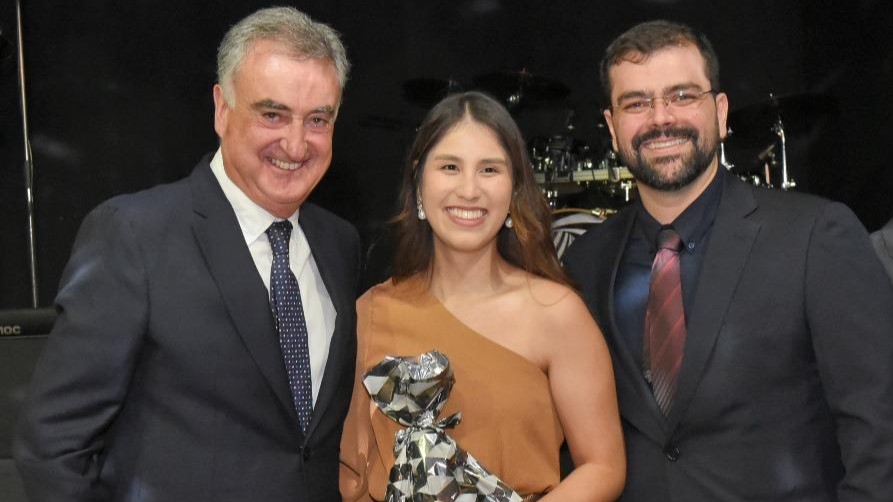 Uma mulher posa sorridente entre dois homens de terno durante cerimônia; ela segura um embrulho prateado.