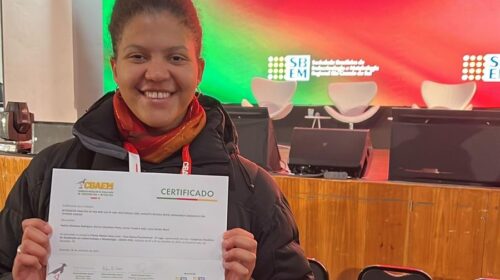 Mulher sorridente segura um certificado com as duas mãos em frente a um palco iluminado com o logotipo do CBAEM 2025. Ela veste casaco preto e cachecol vermelho, e usa um crachá no pescoço.