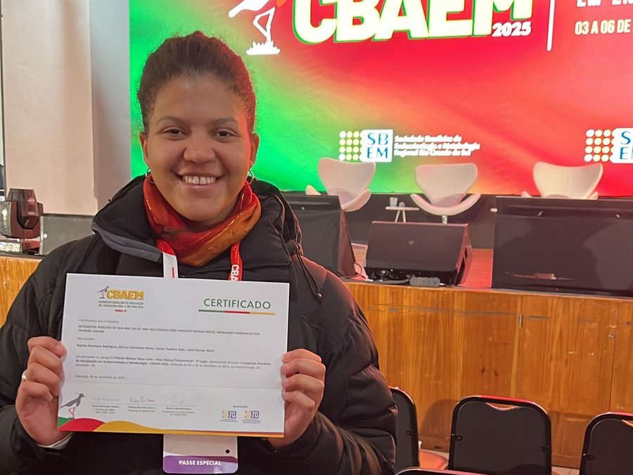 Mulher negra sorridente segura um certificado com as duas mãos em frente a um palco iluminado com o logotipo do CBAEM 2025. Ela veste casaco preto e cachecol vermelho, e usa um crachá no pescoço.