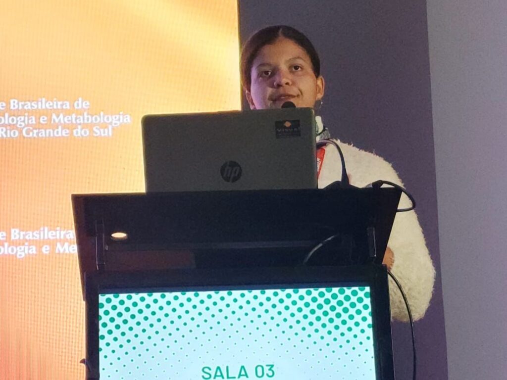 Mulher negra em pé atrás de um púlpito, falando ao microfone durante uma apresentação em congresso. Um notebook HP está à sua frente e no fundo aparece um telão com textos parciais do evento.