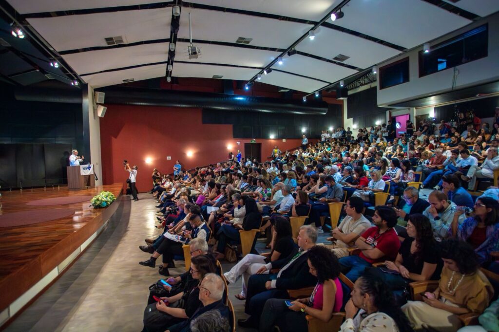 Auditório lotado visto em perspectiva lateral. À esquerda, uma pessoa está em um púlpito com a bandeira do SUS, fazendo uma fala. No palco, há arranjos de flores e uma fotógrafa registrando o momento. À direita, o público ocupa quase todas as cadeiras, atento à apresentação, com algumas pessoas usando celulares ou fazendo anotações. O ambiente é amplo, com teto branco, paredes vermelhas e iluminação difusa.