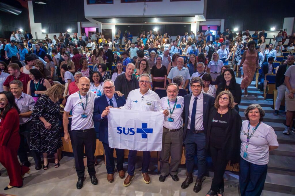 Auditório cheio com pessoas em pé e sentadas, sorrindo e olhando para a câmera. Na frente, um grupo de oito pessoas posa lado a lado, segurando uma bandeira branca com o logotipo do SUS em azul. O ambiente é iluminado, com fileiras de assentos e escadas laterais, transmitindo clima de evento institucional ou celebração.