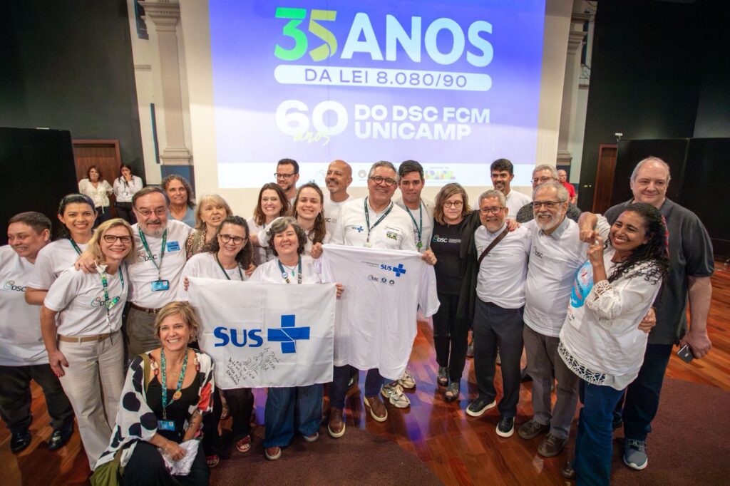 Um grupo de pessoas posa sorridente em frente a uma tela de projeção que exibe os dizeres “35 anos da Lei 8.080/90” e “60 anos do DSC FCM Unicamp”. Algumas pessoas seguram uma camiseta e uma bandeira brancas com o logotipo do SUS em azul. O clima é de celebração e confraternização, com participantes próximos uns dos outros no palco de um auditório.