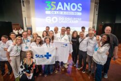 Um grupo de pessoas posa sorridente em frente a uma tela de projeção que exibe os dizeres “35 anos da Lei 8.080/90” e “60 anos do DSC FCM Unicamp”. Algumas pessoas seguram uma camiseta e uma bandeira brancas com o logotipo do SUS em azul. O clima é de celebração e confraternização, com participantes próximos uns dos outros no palco de um auditório.