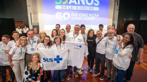 Um grupo de pessoas posa sorridente em frente a uma tela de projeção que exibe os dizeres “35 anos da Lei 8.080/90” e “60 anos do DSC FCM Unicamp”. Algumas pessoas seguram uma camiseta e uma bandeira brancas com o logotipo do SUS em azul. O clima é de celebração e confraternização, com participantes próximos uns dos outros no palco de um auditório.
