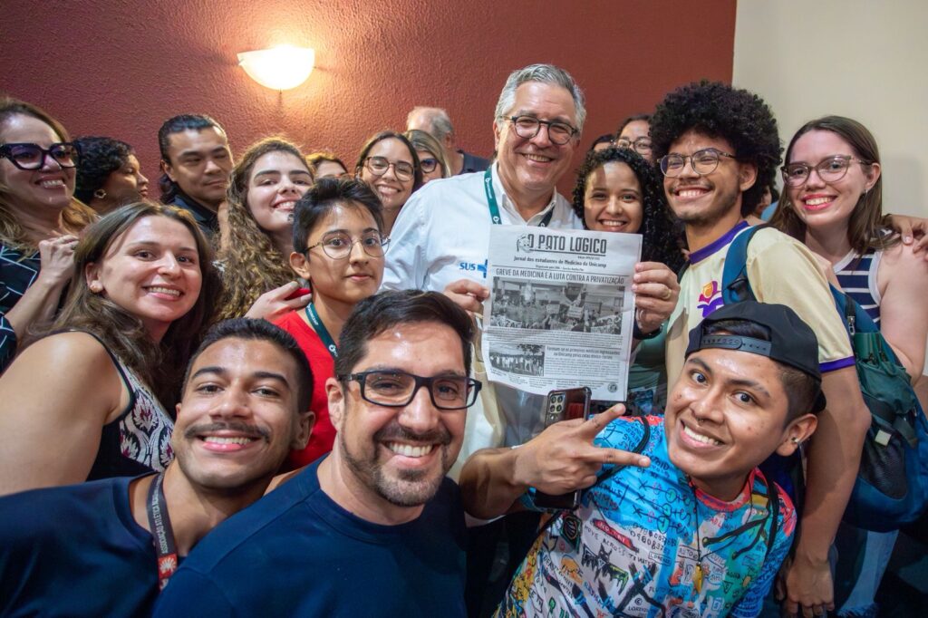 Um grupo de pessoas sorridentes posa para a foto em um ambiente interno, com uma parede vermelha ao fundo. No centro, um homem de óculos e camisa branca segura um jornal intitulado Pato Lógico, que traz como manchete “Greve da Medicina e a Luta contra a Privatização”. Ao redor dele, estudantes de diferentes idades e estilos fazem poses, alguns mostrando o sinal de paz e outros apenas sorrindo para a câmera.