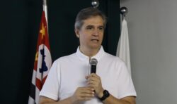 Homem de camisa polo branca fala ao microfone em um púlpito de acrílico. Ao fundo, aparecem parcialmente a bandeira do estado de São Paulo e uma bandeira branca.