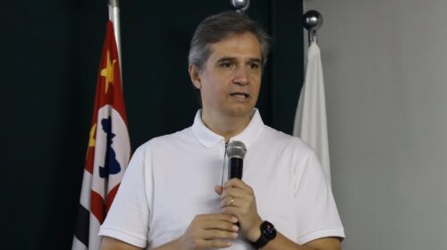 Homem de camisa polo branca fala ao microfone em um púlpito de acrílico. Ao fundo, aparecem parcialmente a bandeira do estado de São Paulo e uma bandeira branca.