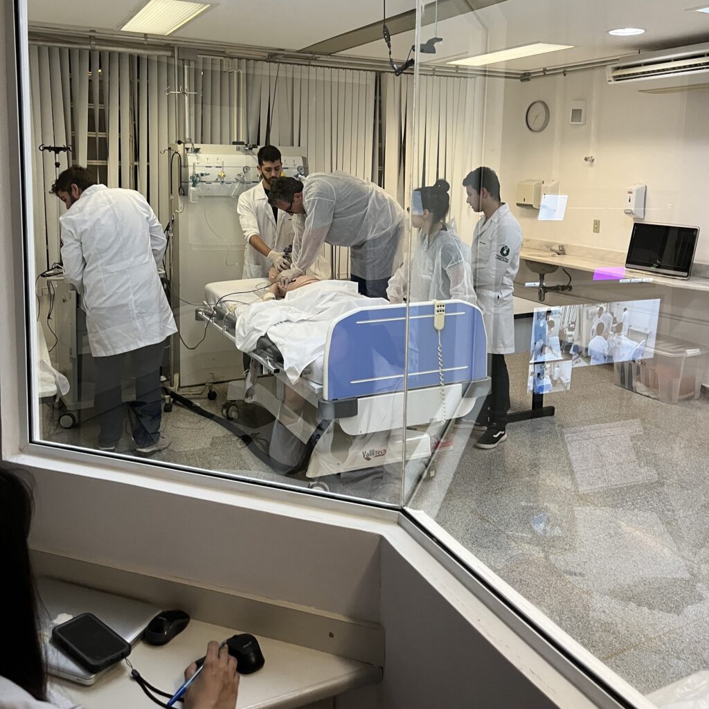 Grupo de estudantes e instrutores de jaleco realizando simulação de atendimento em leito hospitalar, observados através de uma janela de vidro a partir da sala de controle. Ao fundo, equipamentos médicos e monitores