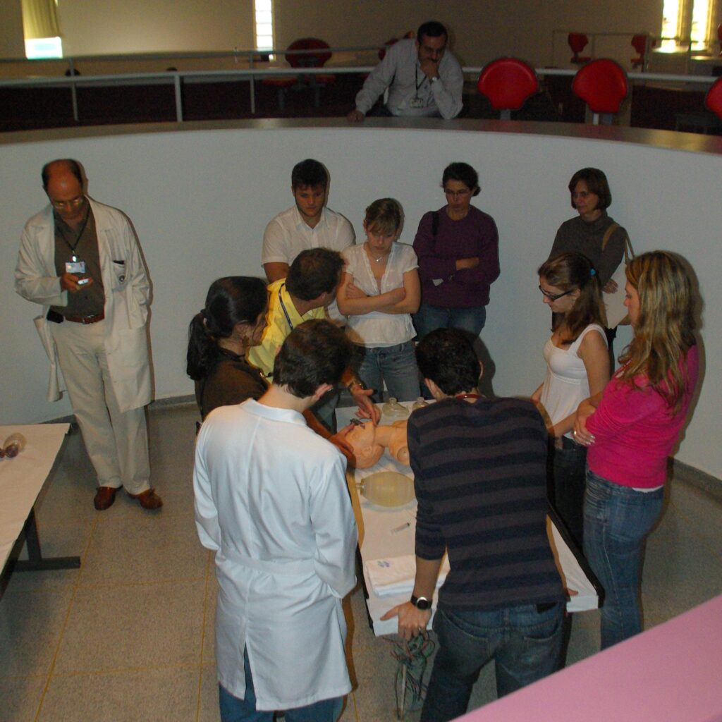 Turma de alunos e professores reunidos ao redor de um manequim de simulação em um espaço circular de ensino, durante atividade prática em ambiente de aprendizado interativo.