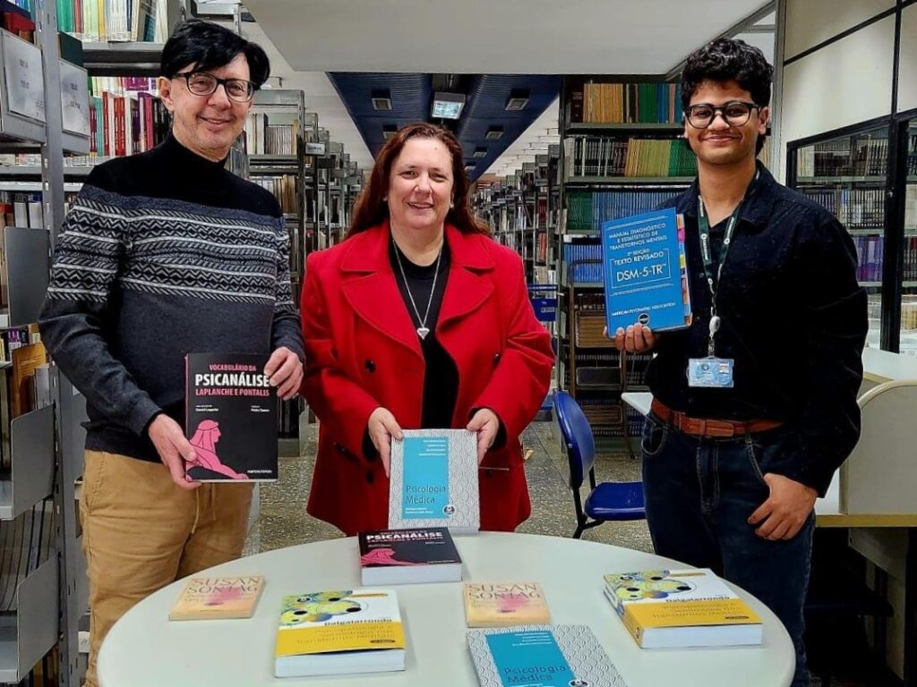 Na biblioteca da FCM, três pessoas estão em pé atrás de uma mesa redonda onde estão expostos vários livros de Psicologia Médica e áreas relacionadas. À esquerda, um homem de suéter cinza e óculos segura o livro Vocabulário da Psicanálise. Ao centro, uma mulher de casaco vermelho segura um exemplar de Psicologia Médica. À direita, um jovem de crachá e camisa escura segura o Manual Diagnóstico e Estatístico de Transtornos Mentais – DSM-5-TR. Ao fundo, há estantes repletas de livros.