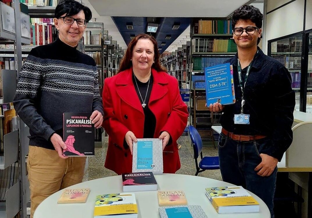 Na biblioteca da FCM, três pessoas estão em pé atrás de uma mesa redonda onde estão expostos vários livros de Psicologia Médica e áreas relacionadas. À esquerda, um homem de suéter cinza e óculos segura o livro Vocabulário da Psicanálise. Ao centro, uma mulher de casaco vermelho segura um exemplar de Psicologia Médica. À direita, um jovem de crachá e camisa escura segura o Manual Diagnóstico e Estatístico de Transtornos Mentais – DSM-5-TR. Ao fundo, há estantes repletas de livros.